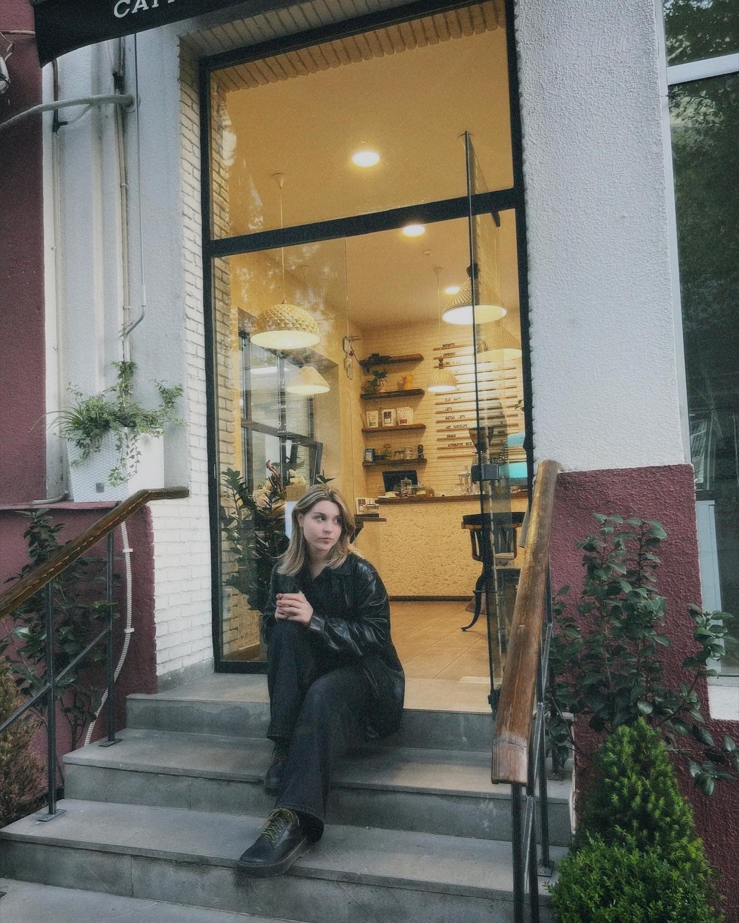 Young woman in a black leather jacket sits on steps outside a warmly lit cafe.