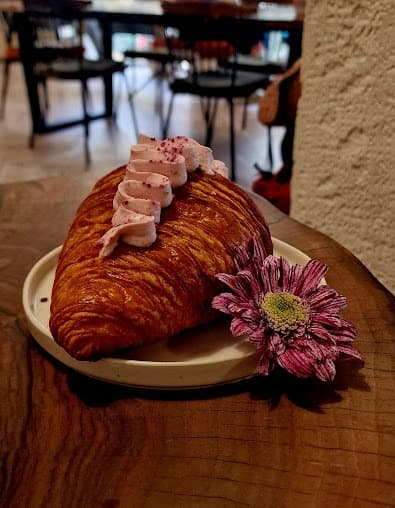 Flaky golden croissant with pink frosting and sprinkles, served alongside a vibrant purple flower.