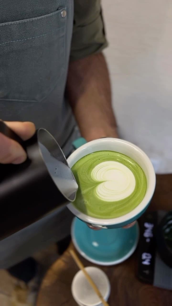 Barista pouring steamed milk into a green matcha latte to create a white heart.