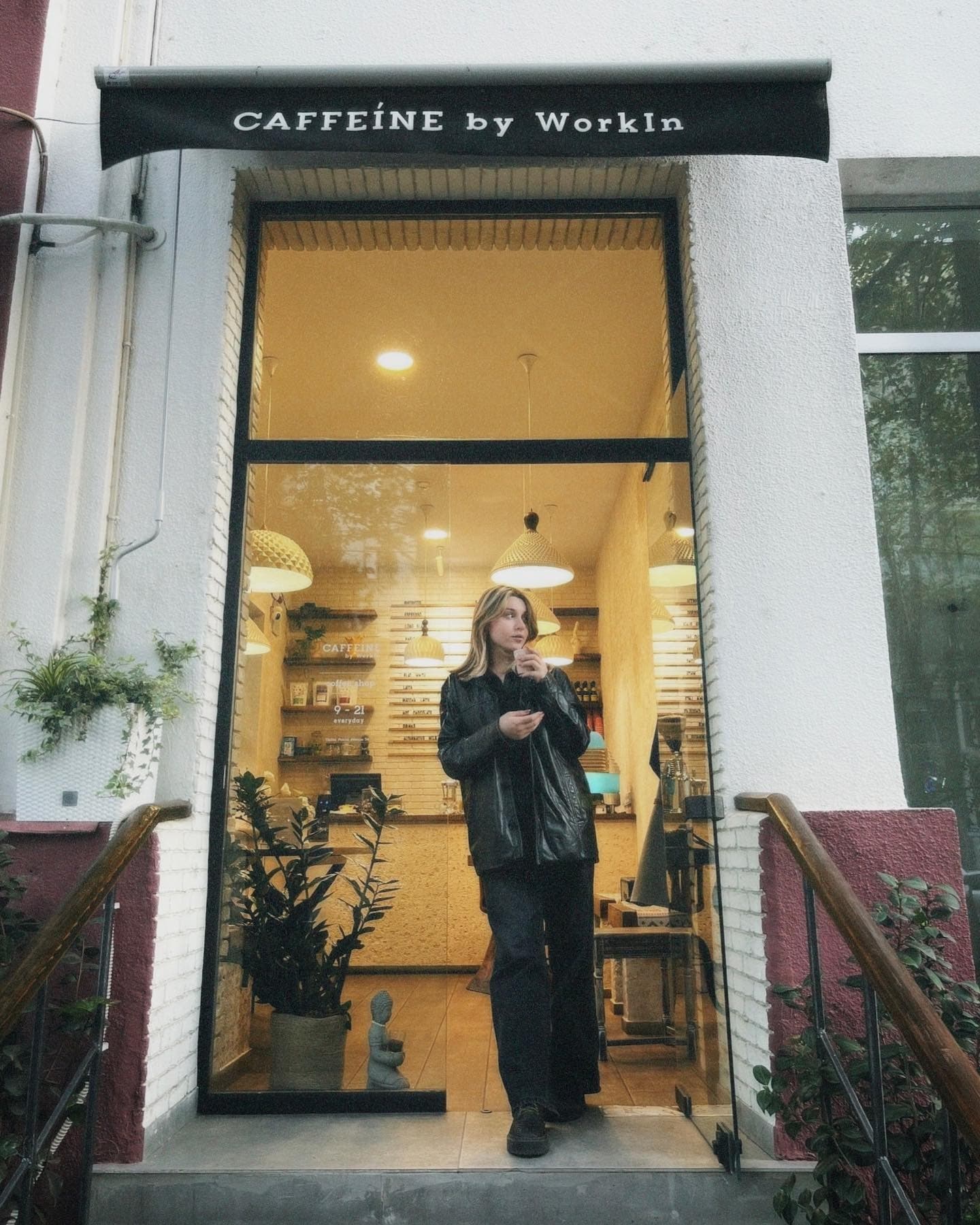 Woman in a leather jacket stands in the doorway of a warmly lit cafe.