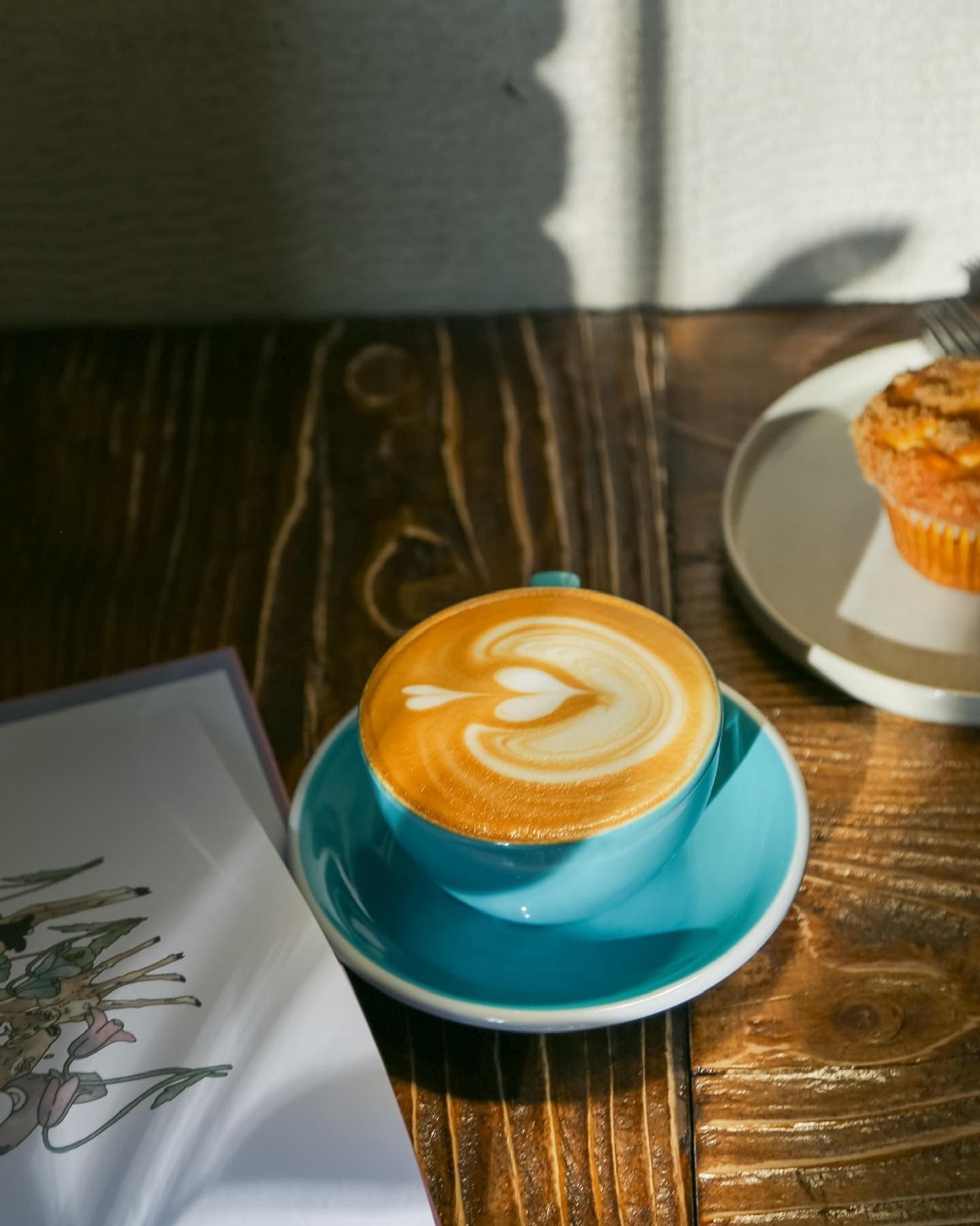 Heart-shaped latte art in a turquoise cup on a rustic wooden table in bright sunlight.