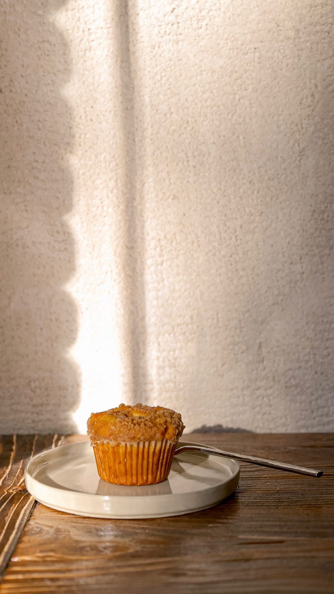 Crumb-topped muffin on a white plate, resting on a wooden table under soft, warm sunlight.