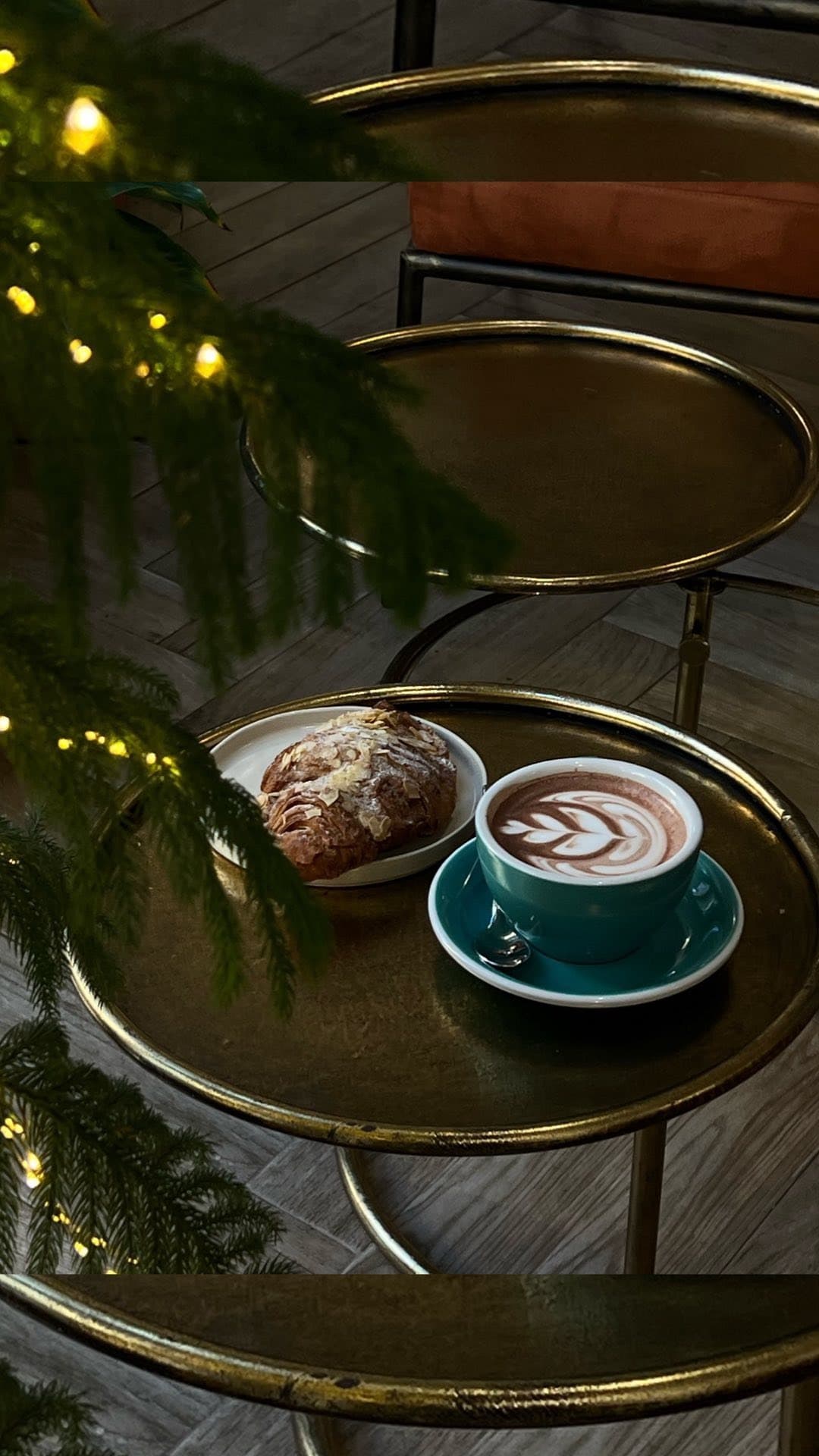 Teal latte cup and almond croissant on a gold table beside a lit Christmas tree.