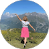 Girl in a pink skirt stands in a mountain meadow with arms outstretched.
