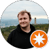 Smiling man in a black hoodie with a scenic city backdrop and orange star icon.
